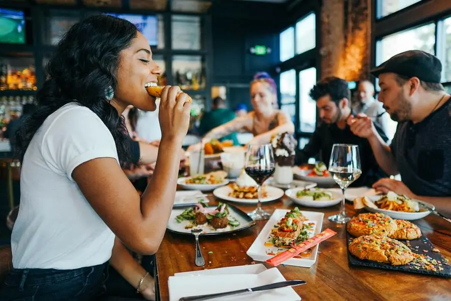 Pessoas comendo e bebendo num restaurante, no primeiro plano uma mulher jovem preta e de fundo dois homens jovens mais uma mulher branca loira