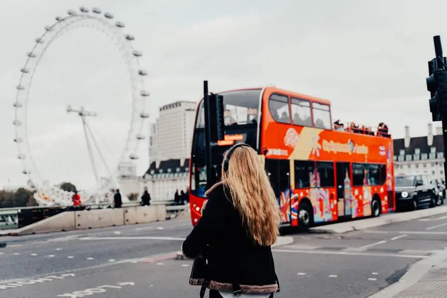 Mulher loira atravesaando a rua com fones de ouvido esperando um ônibus e um carro passarem, no lado esquerdo uma roda gigante