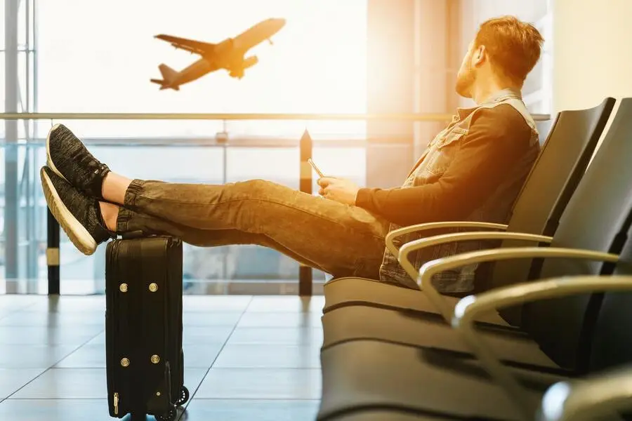 Homem adulto sentado em cadeira num aeroporto com as pernas repousadas mala observa um avião levantando voo