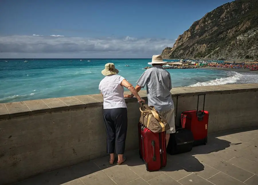 Casal de idosos com malas e chapéu admirando o mar da praia durante o dia. Reprodução Unsplash.