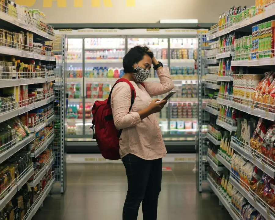 Mulher com celular na mão, mochila vermelha nas costas e máscara no rosto escolhe produtos no supermercado, ela se encontra no corredor de alimentos