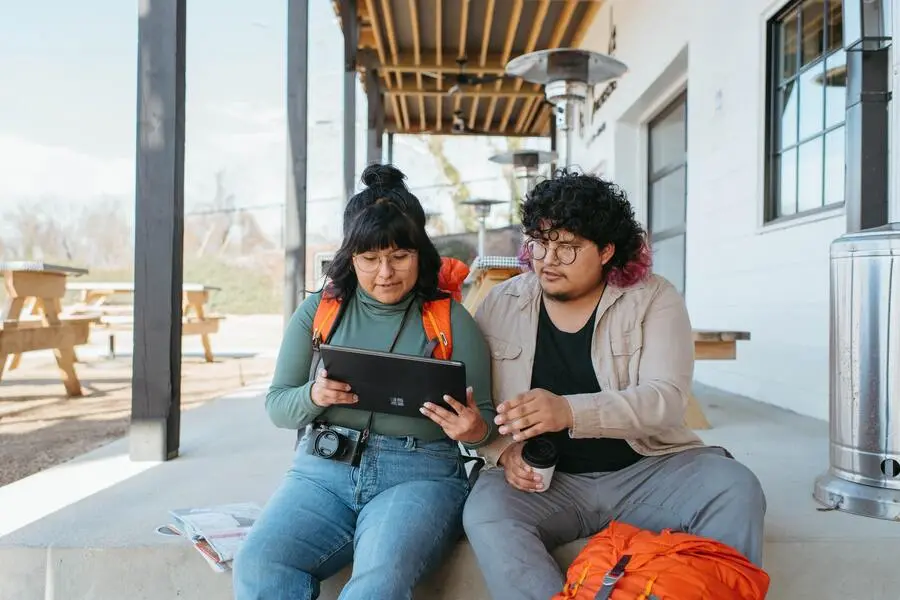 Um casal de amigos vê o planejamento de viagem num tablet, sentados na varanda de uma casa durante o dia